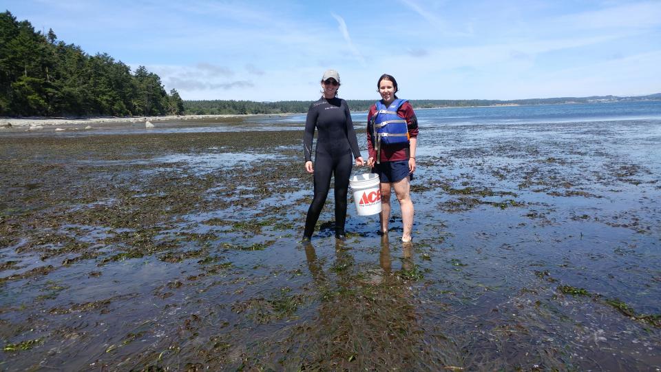 Tori and a labmate hold a bucket in a mud flat